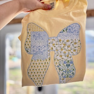 May include: A yellow t-shirt with a large, quilted bow applique. The bow features a light blue center and sections with floral and checkered patterns. The shirt is held up by a hand, and the tag reads "Comfort Colors".