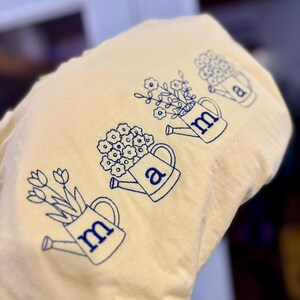 Puede incluir: Una tela amarillo claro con regaderas bordadas en azul, cada una llena de flores. Las letras "m" y "a" est&aacute;n bordadas en las regaderas. El dise&ntilde;o tiene un tema floral.