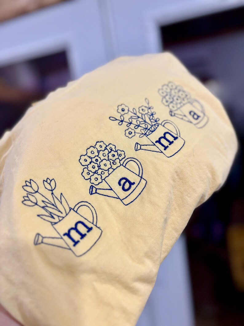 Puede incluir: Una tela amarillo claro con regaderas bordadas en azul, cada una llena de flores. Las letras "m" y "a" est&aacute;n bordadas en las regaderas. El dise&ntilde;o tiene un tema floral.