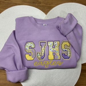 May include: A lavender sweatshirt with the letters "SJHS" in a floral and yellow design. The word "eagles" is embroidered in yellow script below the letters. The sweatshirt is folded on a white, heart-shaped placemat.
