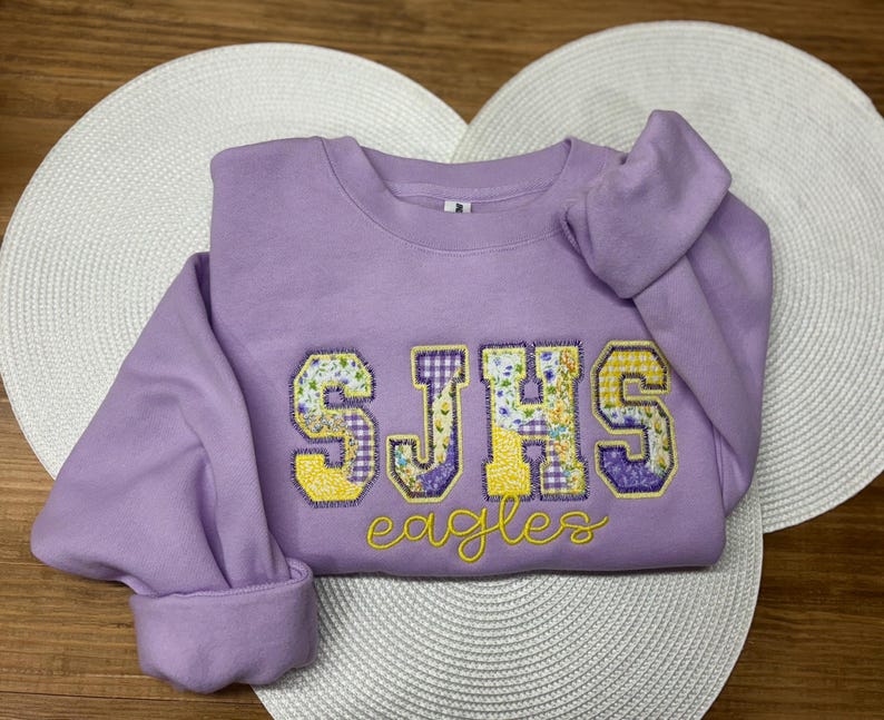 May include: A lavender sweatshirt with the letters "SJHS" in a floral and yellow design. The word "eagles" is embroidered in yellow script below the letters. The sweatshirt is folded on a white, heart-shaped placemat.