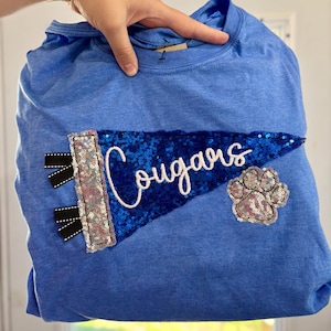 May include: A blue crewneck t-shirt featuring a large, dark blue sequined pennant patch with the word "Cougars" embroidered in white script. The pennant includes a silver sequined border with black ribbon accents and a silver sequined paw print.