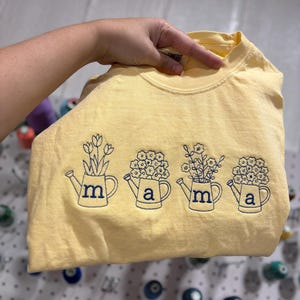 Puede incluir: Camiseta amarilla clara con regaderas bordadas, cada una con una flor y las letras "m", "a", "m" y "a". El dise&ntilde;o es azul marino. La camiseta es de color amarillo claro.