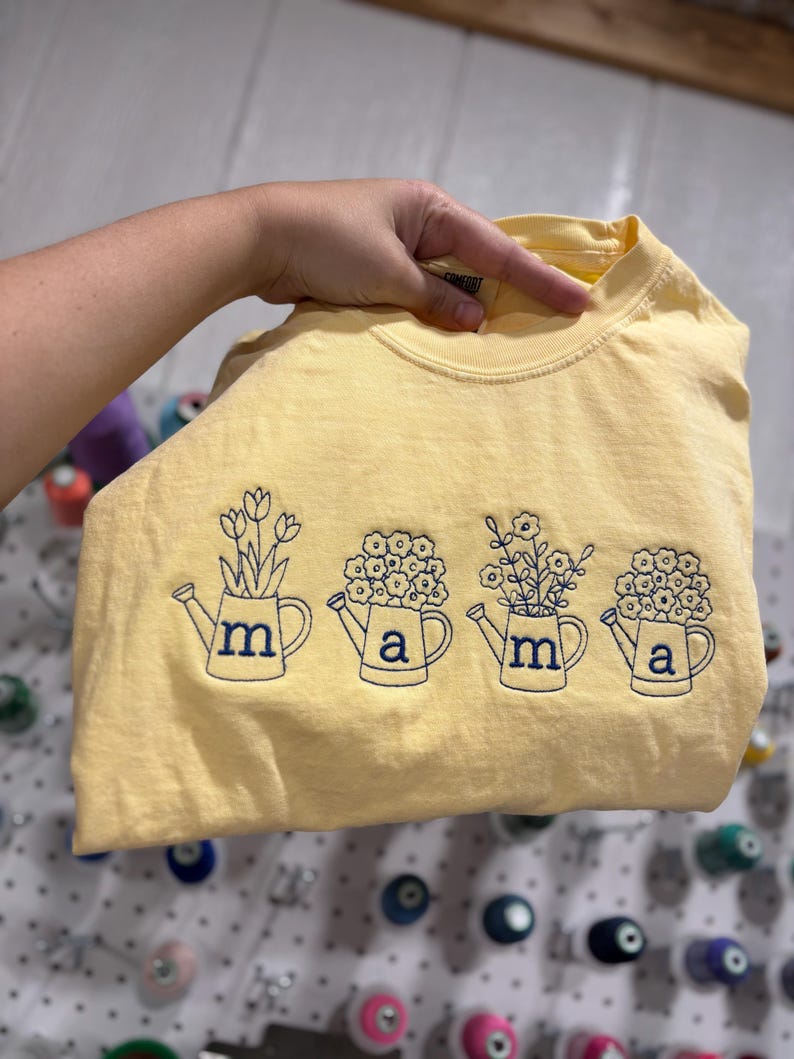 Puede incluir: Camiseta amarilla clara con regaderas bordadas, cada una con una flor y las letras "m", "a", "m" y "a". El dise&ntilde;o es azul marino. La camiseta es de color amarillo claro.
