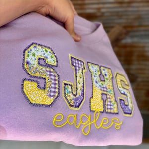 May include: A light purple sweatshirt with the letters "SJHS" in a patchwork design, featuring floral and gingham patterns. The word "eagles" is embroidered in yellow script below the letters. The sweatshirt is held up by a hand.