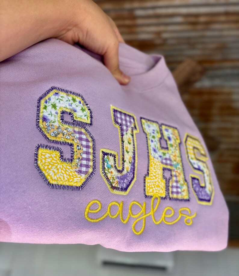 May include: A light purple sweatshirt with the letters "SJHS" in a patchwork design, featuring floral and gingham patterns. The word "eagles" is embroidered in yellow script below the letters. The sweatshirt is held up by a hand.