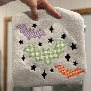 May include: A light gray sweatshirt featuring an embroidered design of three bats in purple, green, and orange gingham patterns. The design is accented with several small, solid black embroidered stars scattered around the bats.