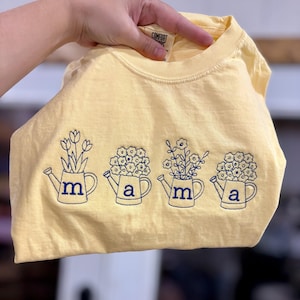 Puede incluir: Una camiseta amarilla clara con un dise&ntilde;o floral azul marino. El dise&ntilde;o presenta cuatro regaderas con las letras "m" y "a". Cada regadera tiene un arreglo floral diferente. La camiseta est&aacute; hecha de un material suave.