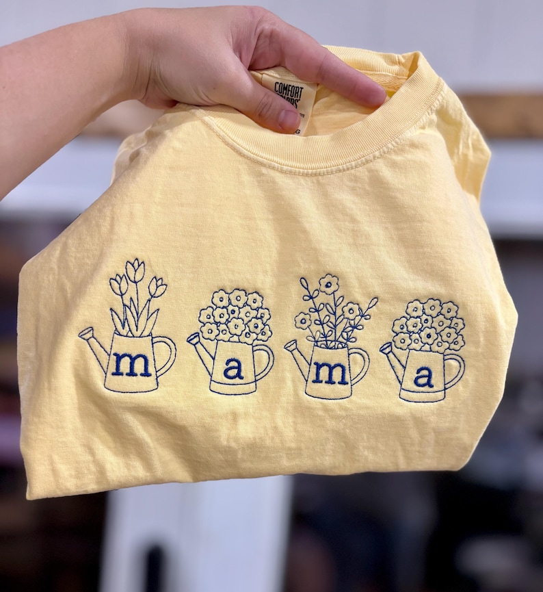 Puede incluir: Una camiseta amarilla clara con un dise&ntilde;o floral azul marino. El dise&ntilde;o presenta cuatro regaderas con las letras "m" y "a". Cada regadera tiene un arreglo floral diferente. La camiseta est&aacute; hecha de un material suave.