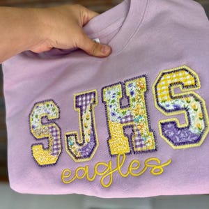 May include: A light purple sweatshirt with the letters "SJHS" appliqued in a patchwork design of floral and gingham fabrics. The word "eagles" is embroidered in yellow script below the letters.