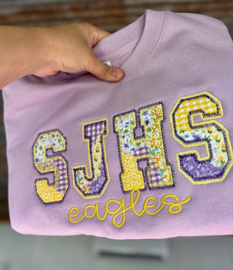 May include: A light purple sweatshirt with the letters "SJHS" appliqued in a patchwork design of floral and gingham fabrics. The word "eagles" is embroidered in yellow script below the letters.