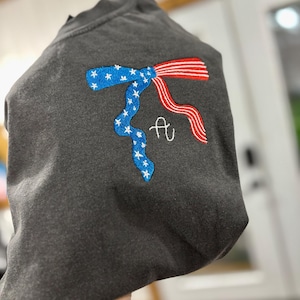 May include: A charcoal gray sweatshirt featuring an embroidered design of a ribbon tied in a bow. One side of the ribbon is blue with white stars, and the other side features red and white stripes. A white cursive letter A is embroidered below the bow.