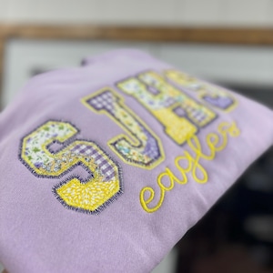May include: A light purple hoodie with the letters "SJH" in a patchwork design of floral, gingham, and yellow patterns. The word "Eagles" is embroidered in yellow script below the letters.