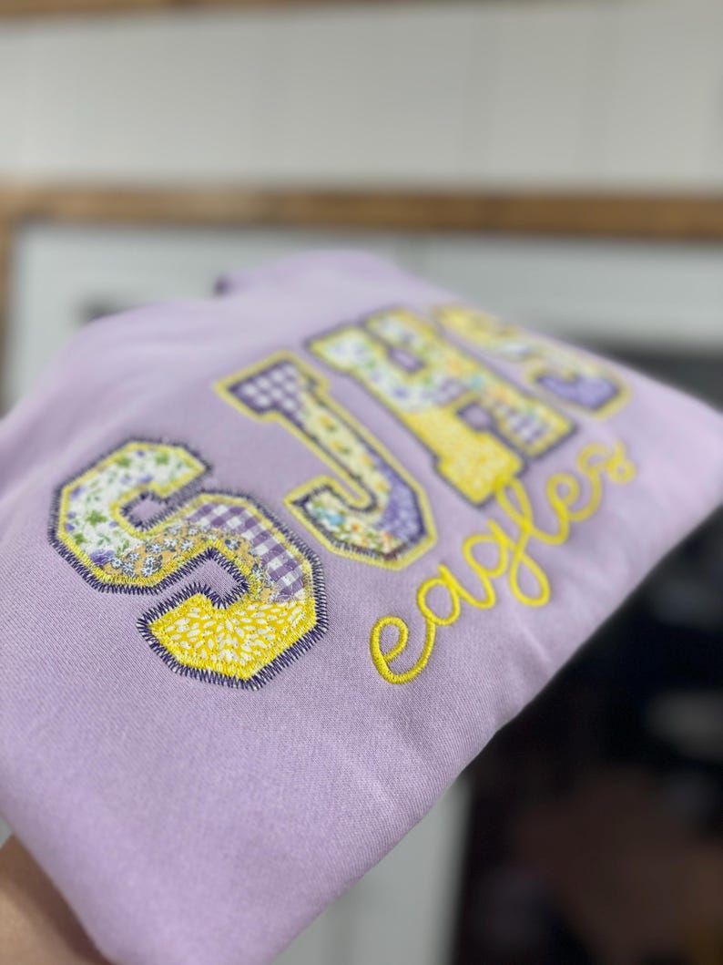 May include: A light purple hoodie with the letters "SJH" in a patchwork design of floral, gingham, and yellow patterns. The word "Eagles" is embroidered in yellow script below the letters.