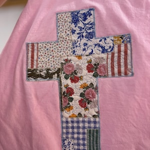 May include: A light pink sweatshirt with a patchwork cross design. The cross is made of various fabric patterns, including floral, striped, and checkered designs, in shades of blue, red, and green.