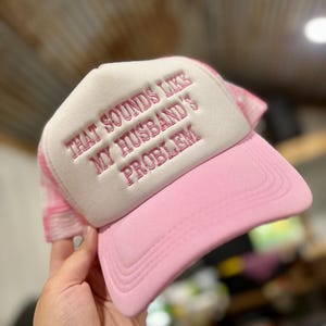 May include: Pink and white trucker hat with the embroidered text "THAT SOUNDS LIKE MY HUSBAND'S PROBLEM". The hat has a pink brim and a white front panel with the text in pink. The back of the hat is a pink and white mesh.