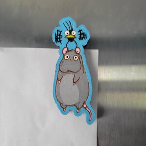 May include: A wooden magnet featuring a gray mouse with a blue bird perched on its head. The mouse has pink ears and a long tail. The bird has black eyes and a yellow beak.