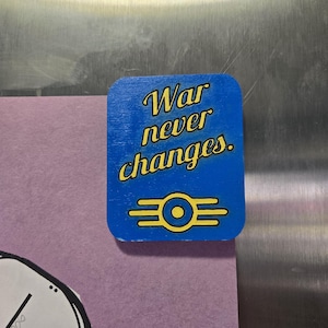 May include: A blue, rectangular magnet with the text "War never changes." in yellow script. Below the text is a yellow symbol with a circle in the center and two horizontal lines on each side. The magnet is on a metal surface.