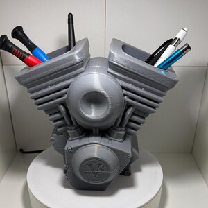 Könnte beinhalten: Ein grauer, 3D-gedruckter Schreibtisch-Organizer in Form eines V-Twin-Motors. Der Organizer hat zwei Fächer mit Stiften und Werkzeugen mit roten, blauen, schwarzen und weißen Akzenten. Die Basis ist eine weiße, kreisförmige Plattform.