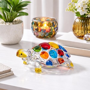 May include: A colorful glass turtle figurine with a mosaic shell of red, yellow, blue, and green. The turtle is on a white surface, with a candle holder and a small vase with flowers in the background. The figurine is approximately 6 inches long.