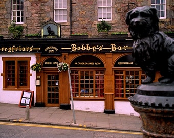 May include: A black and white sign with gold lettering reads "Greyfriars" and "Bobby's Bar" with an arrow pointing to the right. The sign is above a pub with a brown door and windows. The pub is located on a street with a cobblestone sidewalk. A black statue of a dog sits on a pedestal in front of the pub.