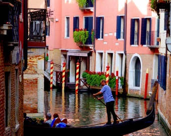 May include: A gondola with two people in it is being rowed down a canal in Venice, Italy. The gondola is black and has a red stripe on the side. The canal is lined with colorful buildings. The text "Splendido Venica" is visible on the building.