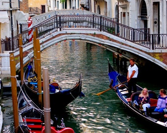 May include: Three gondolas are docked in a canal in Venice, Italy. The gondolas are black with red and blue accents. The gondolas are in the foreground of the image, with a bridge and buildings in the background.