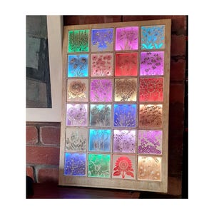 May include: A wooden framed light box with 25 illuminated squares, each featuring a different floral design. The lights cycle through various colours, creating a vibrant display. The frame is a light wood colour, and the overall effect is decorative.