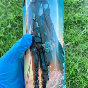 May include: A stainless steel tumbler featuring a colourful image of a character from the Star Wars series, The Mandalorian, and a small green creature, Grogu, walking on a desert planet. The tumbler has a green Starbucks logo on the bottom.