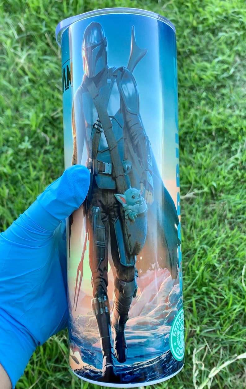 May include: A stainless steel tumbler featuring a colourful image of a character from the Star Wars series, The Mandalorian, and a small green creature, Grogu, walking on a desert planet. The tumbler has a green Starbucks logo on the bottom.