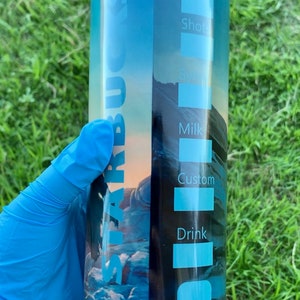 May include: A blue and white stainless steel tumbler with a Starbucks logo and a mountain scene. The tumbler has a list of drink options on the side, including decaf, shots, syrup, milk, custom, and drink.