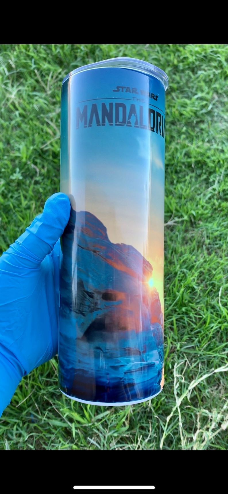 May include: A blue and orange tumbler with a white lid. The tumbler features a graphic of a sunset over a rocky landscape and the text "Star Wars The Mandalorian".