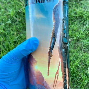 May include: A stainless steel tumbler featuring a blue, green and orange graphic of a character from the Star Wars series, The Mandalorian. The text "The Mandalorian" is printed on the tumbler.
