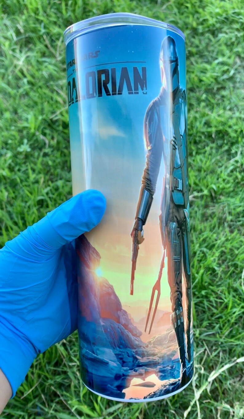 May include: A stainless steel tumbler featuring a blue, green and orange graphic of a character from the Star Wars series, The Mandalorian. The text "The Mandalorian" is printed on the tumbler.