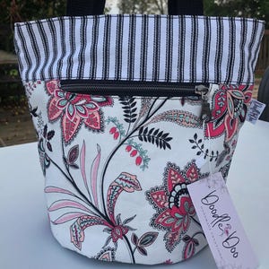 May include: A small tote bag with a black and white striped top and a floral pattern on the body. The floral pattern features pink and red flowers and leaves on a white background. The bag has a black zipper and black handles.
