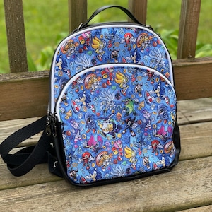 Puede incluir: Una mochila azul con un patrón colorido de personajes de Sonic the Hedgehog. La mochila tiene una base negra, una cremallera blanca y un asa y correas negras. El diseño incluye varios personajes y elementos del universo Sonic.