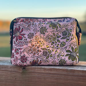 May include: A pink zippered pouch with a floral print featuring black, green, and red flowers. The pouch is made of a soft, water-resistant material.