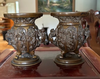 May include: Two antique bronze vases with intricate relief carvings of mythical creatures and floral patterns. Each vase has a wide, flat top, a bulbous body, and three small feet. The vases are displayed on a dark wooden surface.