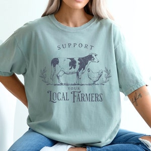 May include: A light blue t-shirt with a vintage-style graphic of a cow and a chicken. The graphic says "Support Your Local Farmers".