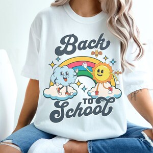 May include: A white t-shirt with a retro-style graphic design featuring a smiling sun and a cloud holding hands over a rainbow. The text 'Back to School' is written in a retro font below the graphic.