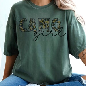May include: A green t-shirt with the words "CAMO girl" printed on it. The letters are outlined in black and filled with a camouflage pattern.