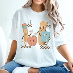 May include: White t-shirt with a graphic of a pencil, ruler, apple, and book with cartoon faces. The graphic is in a retro style with checkered patterns.