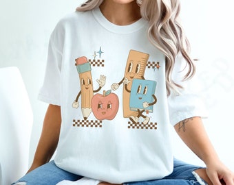 May include: White t-shirt with a graphic of a pencil, ruler, apple, and book with cartoon faces. The graphic is in a retro style with checkered patterns.