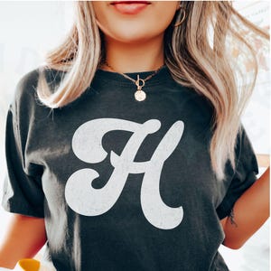 May include: A dark gray t-shirt with a white stylized letter "H" printed on the front. The shirt has a relaxed fit and short sleeves.