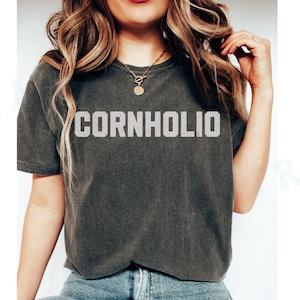 May include: A dark gray t-shirt with the text "CORNHOLIO" printed in white on the front. The shirt is worn by a person with long blonde hair and a gold necklace.