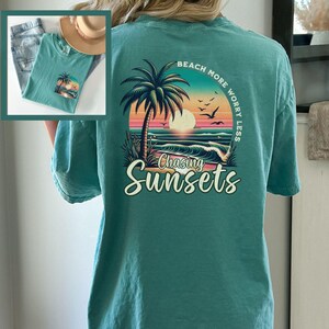 May include: Teal blue t-shirt with a graphic print of a palm tree, sunset, and ocean waves. The text "Beach More Worry Less" is above the graphic and "Chasing Sunsets" is below the graphic.