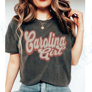 May include: A dark grey t-shirt with a vintage-style graphic that says "Carolina Girl" in red and white lettering.