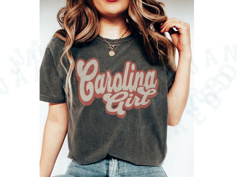 May include: A dark grey t-shirt with a vintage-style graphic that says "Carolina Girl" in red and white lettering.