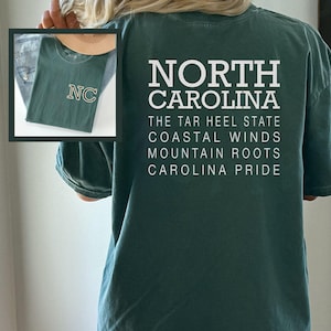 May include: Dark green t-shirt with the words "NORTH CAROLINA" and other text in white. The shirt also features a smaller image of a folded green t-shirt with "NC" on it. The shirt is a casual style.