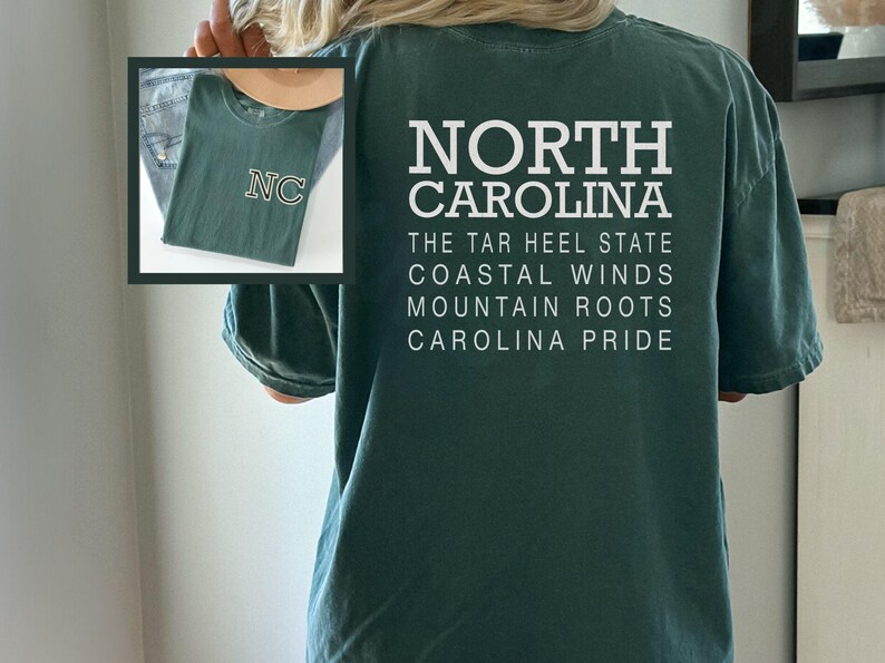 May include: Dark green t-shirt with the words "NORTH CAROLINA" and other text in white. The shirt also features a smaller image of a folded green t-shirt with "NC" on it. The shirt is a casual style.